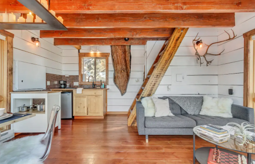 Cozy interior of a cabin with wooden beams, a sofa, kitchenette, and rustic decor, featuring a staircase and antlers.