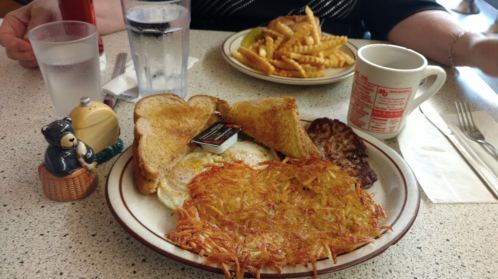 A plate of breakfast with eggs, hash browns, toast, sausage, and a side of fries, with drinks in the background.