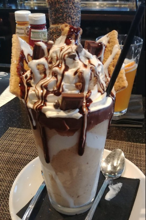 A tall milkshake topped with whipped cream, chocolate drizzle, and cookie pieces, served with a spoon and straw.