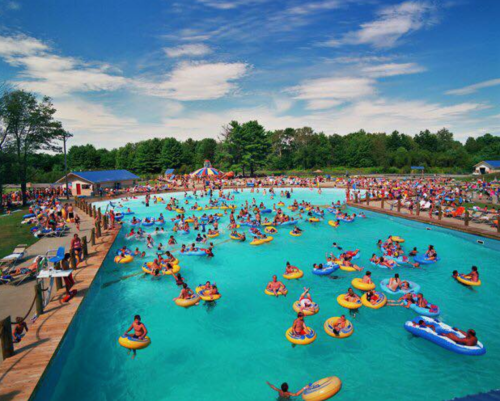 A crowded outdoor pool filled with people on floaties, surrounded by trees and a clear blue sky.