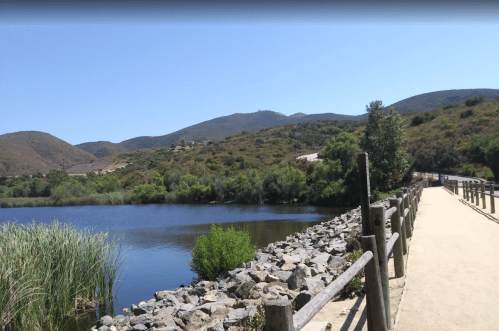 A scenic pathway alongside a calm lake, surrounded by lush greenery and rolling hills under a clear blue sky.