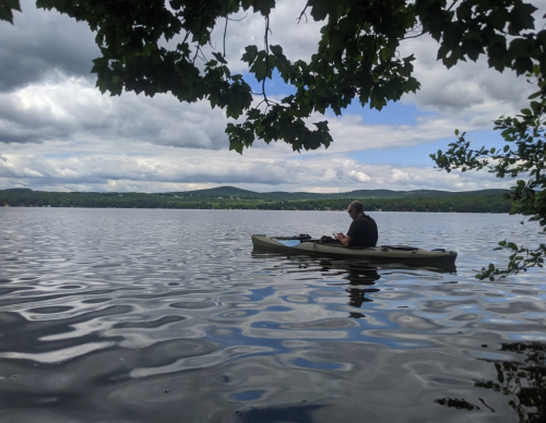 A person sits in a kayak on a calm lake, surrounded by trees and mountains under a cloudy sky.