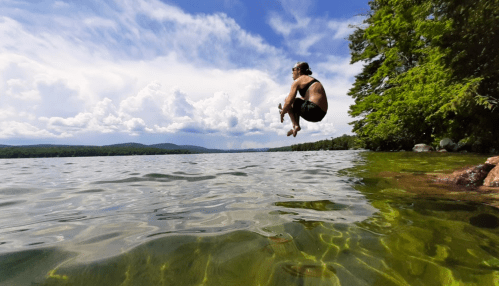 A person jumps into a clear lake on a sunny day, surrounded by trees and mountains under a blue sky with clouds.