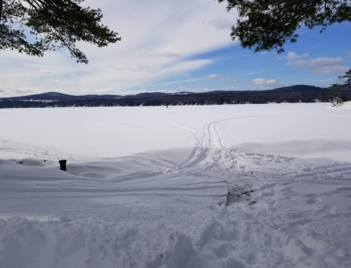 A snowy landscape with a frozen lake, tire tracks leading into the distance, and mountains under a partly cloudy sky.