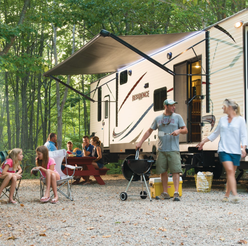 A family gathers around a camper in a wooded area, enjoying a barbecue and socializing outdoors.