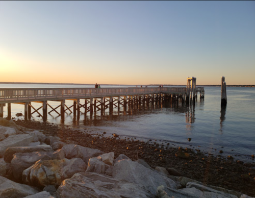 A wooden pier extends into calm water at sunset, with rocks in the foreground and a clear sky.