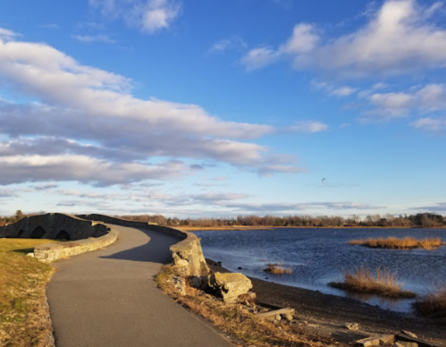 A winding path along a river with a stone bridge, under a blue sky with scattered clouds and grassy banks.