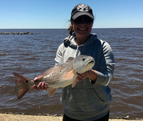 A person in a gray hoodie holds a large fish by the water, with a clear blue sky in the background.
