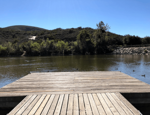 A wooden dock extends over a calm lake, surrounded by green hills and trees under a clear blue sky. Ducks swim nearby.