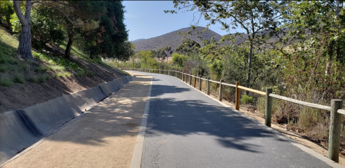 A winding path through greenery, with a dirt trail beside a paved road, surrounded by trees and hills in the background.