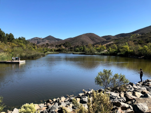 A serene lake surrounded by hills, with people fishing and a small dock on a clear, sunny day.