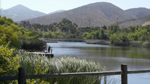 A serene landscape featuring a calm river, lush greenery, and people fishing from a wooden dock with mountains in the background.
