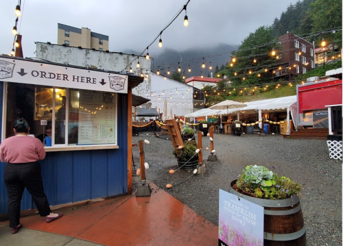 A cozy outdoor market with string lights, food stalls, and a misty mountain backdrop. A person stands at a food order window.