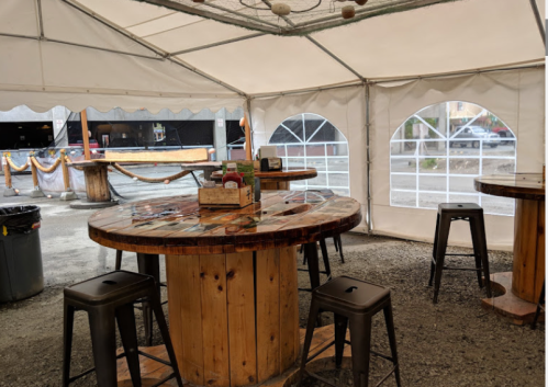 A tented outdoor seating area with wooden tables and metal stools, set up for dining.