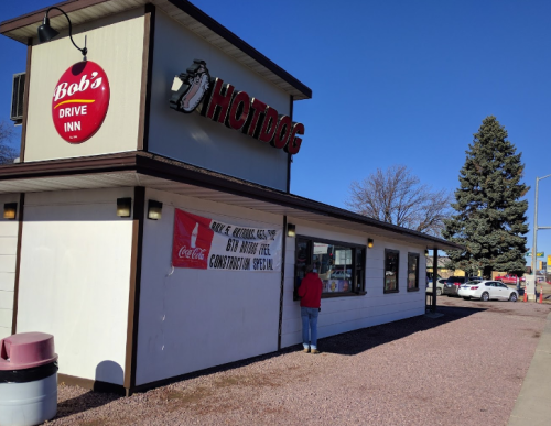 A drive-in restaurant named "Bob's Hotdog" with a customer at the window and a clear blue sky in the background.