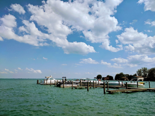 A serene lake scene with boats docked at a wooden pier under a blue sky with fluffy clouds.