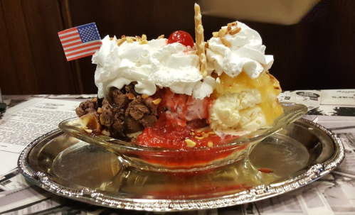 A colorful ice cream sundae topped with whipped cream, cherries, nuts, and a small American flag, served in a silver dish.