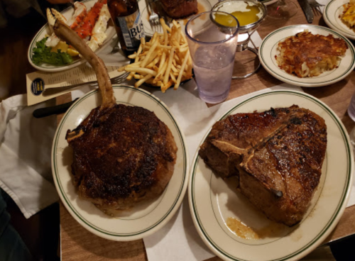 Two large, grilled steaks on plates, accompanied by fries and a side of condiments, set on a wooden table.