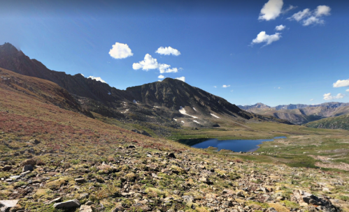 A scenic mountain landscape featuring a clear lake, rocky terrain, and vibrant green and red vegetation under a blue sky.
