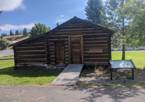 A rustic log cabin with a wooden walkway, surrounded by trees and grass, featuring an informational sign nearby.