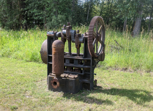 An old, rusted industrial machine with gears and a cylinder, set in a grassy area surrounded by trees.