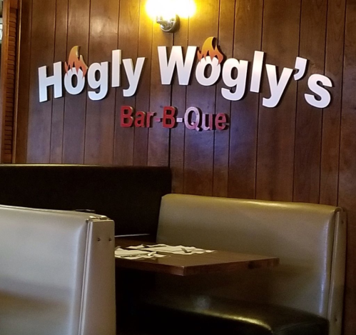 Sign for Hogly Wogly's Bar-B-Que on a wooden wall, with a booth and table in the foreground.