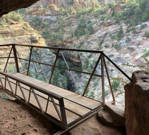 View from a cave opening overlooking a rocky landscape with greenery and a wooden railing in the foreground.