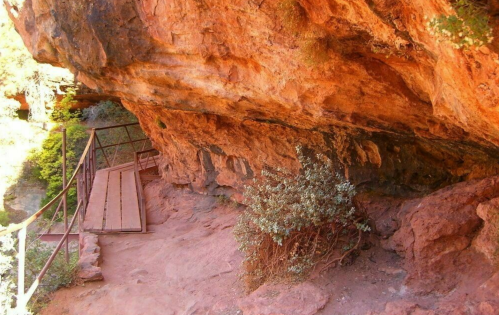 A rocky cliff with a wooden walkway and sparse vegetation beneath a natural overhang.