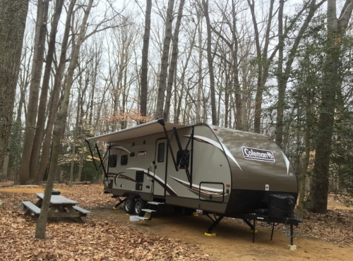 A Coleman travel trailer parked in a wooded area with bare trees and a picnic table nearby.