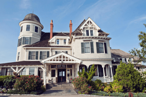 A large, elegant Victorian-style house with a turret, surrounded by lush greenery and colorful flowers.