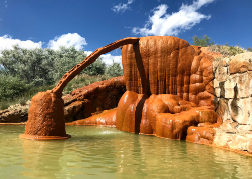 A natural hot spring with orange mineral deposits, featuring a rock formation and a small waterfall into a green pool.