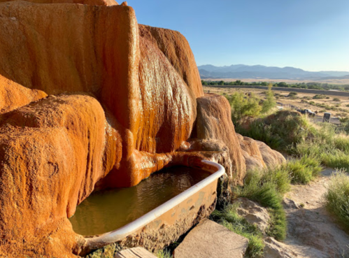Natural hot spring with orange mineral deposits, surrounded by grass and mountains in the background under a clear sky.