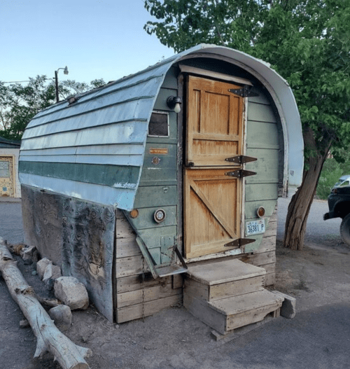 A vintage trailer with a curved roof, wooden door, and steps, surrounded by trees and gravel.