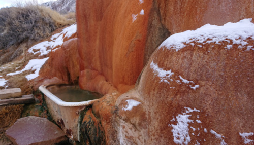 A rustic, weathered bathtub sits against a reddish rock wall, partially covered in snow.