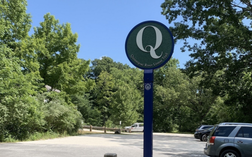Sign for the Quinebaug River Linear Park, surrounded by greenery and a parking area with parked cars.