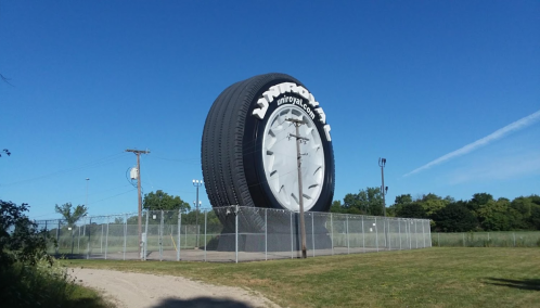 Giant tire sculpture on a fenced lot under a clear blue sky, surrounded by grass and trees.