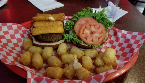 A burger with lettuce, tomato, and cheese, served with crispy tater tots in a red basket with checkered paper.