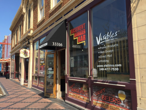 Exterior view of a building featuring Basement Burger Bar and Visibles Marketing, with signage and a brick walkway.