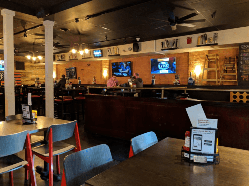 A cozy bar interior with wooden accents, tables, and a long counter, featuring TVs and patrons enjoying drinks.