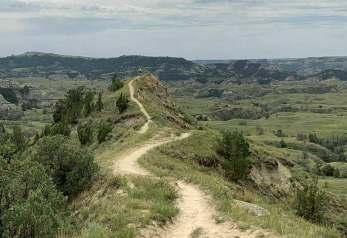 A winding dirt path leads up a grassy hill, surrounded by rolling green landscapes and distant hills under a cloudy sky.