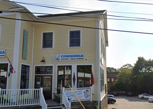 A yellow building with a sign for "Cowabunga Smoothies & Frozen Lemonade," featuring a porch and nearby parking.