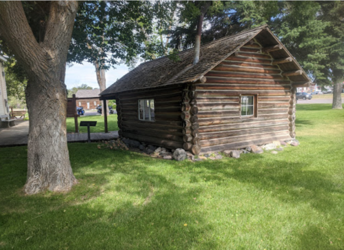 A small log cabin surrounded by grass and trees, with a sign nearby in a park setting.