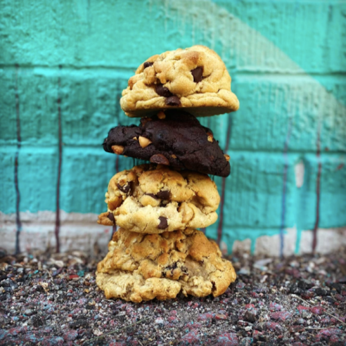 A stack of four cookies, including chocolate chip and chocolate varieties, against a vibrant turquoise wall.