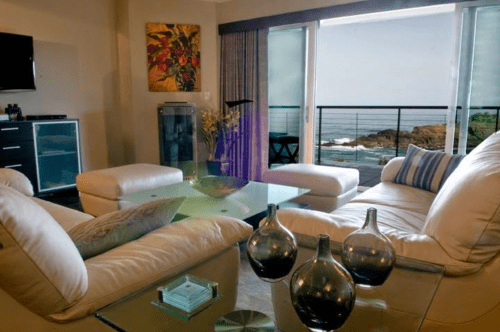 Modern living room with white furniture, glass table, and ocean view through large windows.
