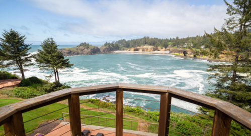 Scenic view of a coastal landscape with waves, trees, and a cloudy sky from a wooden balcony.
