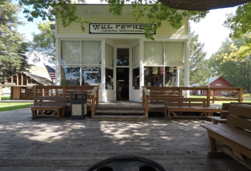 A quaint general merchandise store with large windows, surrounded by trees and wooden benches on a sunny day.