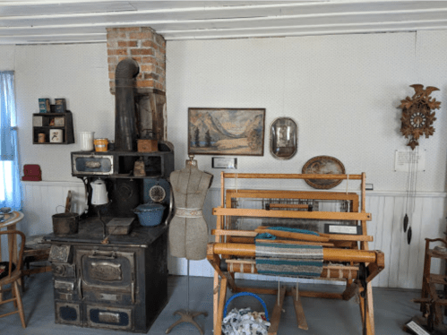 A vintage room featuring a wood stove, loom, antique decor, and a dress form against a patterned wall.