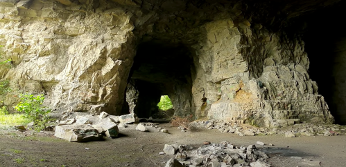A rocky cave interior with large stone walls and scattered boulders, illuminated by natural light from the entrance.