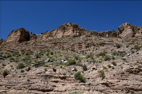 A rocky desert landscape with layered cliffs under a clear blue sky, featuring sparse vegetation and rugged terrain.