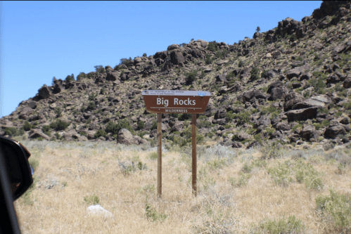 Sign for "Big Rocks Wilderness" in a grassy area with rocky hills in the background under a clear blue sky.